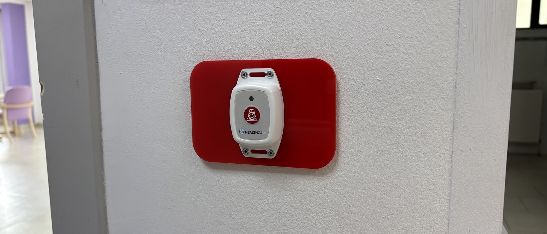 Bouton d'appel sans fil Bluetooth Healthcall fixé sur une plaque de finition acrylique rouge, qui masque l'emplacement de l'ancien bouton Télévic, sur un mur blanc de couloir