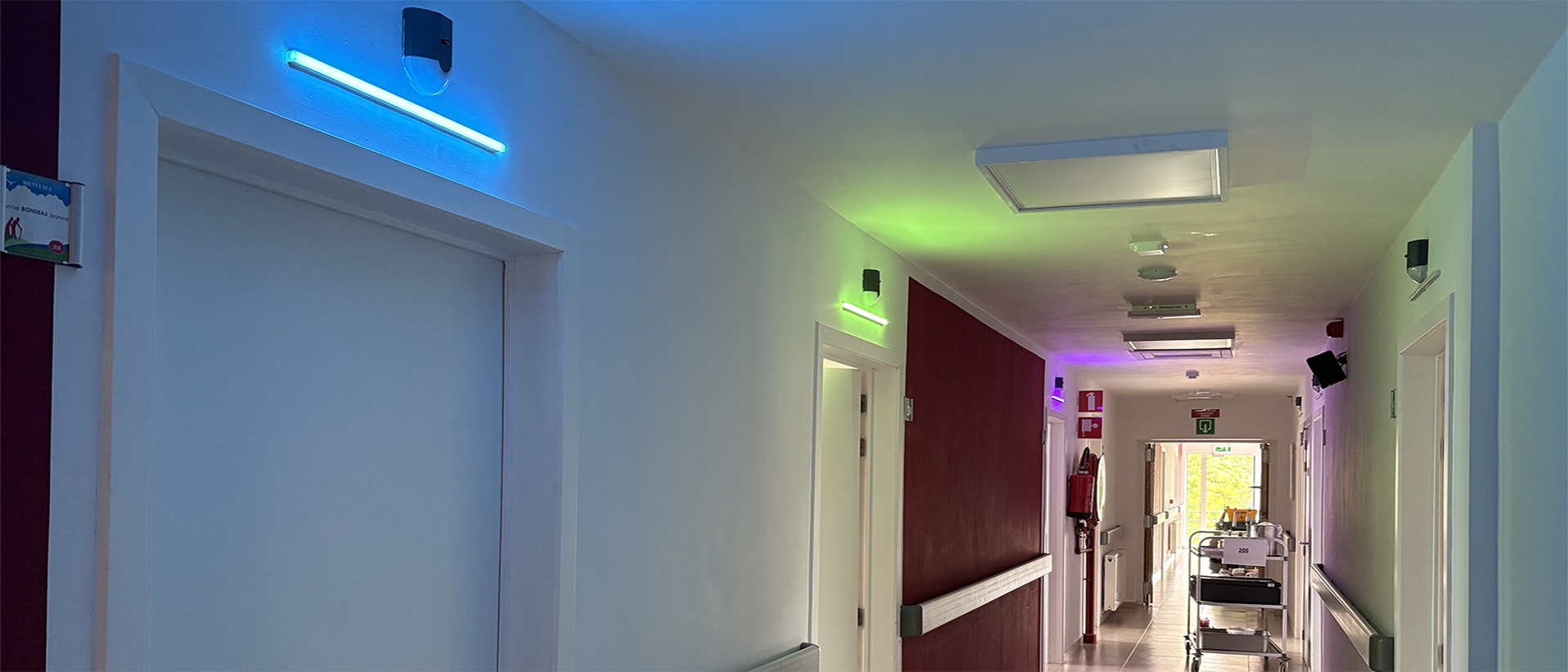 Couloir d'étage de la résidence Le Petit Paradis vu de nuit, hublots LED de 50 cm au-dessus de chaque porte de chambre en cyan, vert et magenta, ambiance calme et veillée