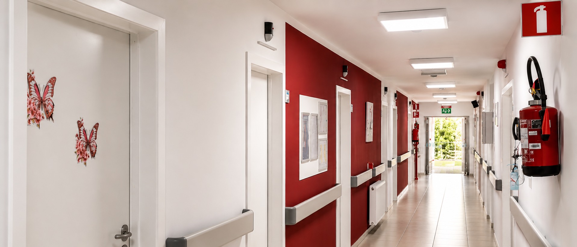 Couloir d'étage de la résidence Le Petit Paradis, murs blancs et accents rouges, hublots LED Healthcall au plafond, lumière naturelle au fond du couloir