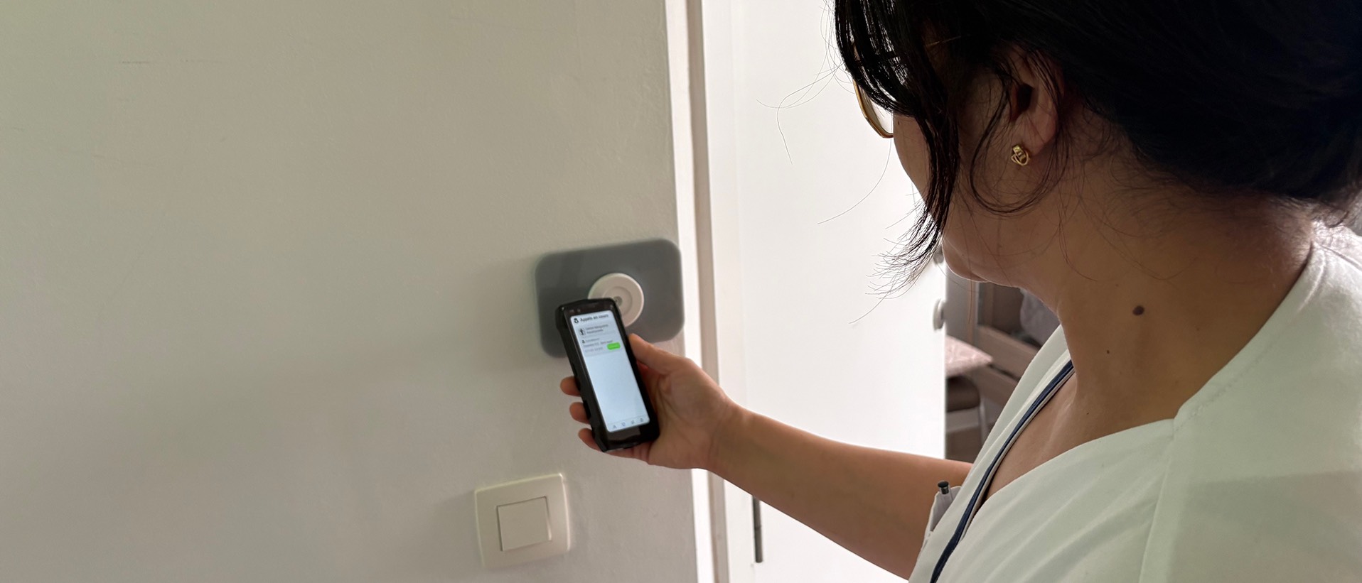 Soignante en tenue claire face à l'entrée d'une chambre, smartphone industriel 4,5 pouces en main affichant l'application Healthcall, à côté du lecteur NFC mural