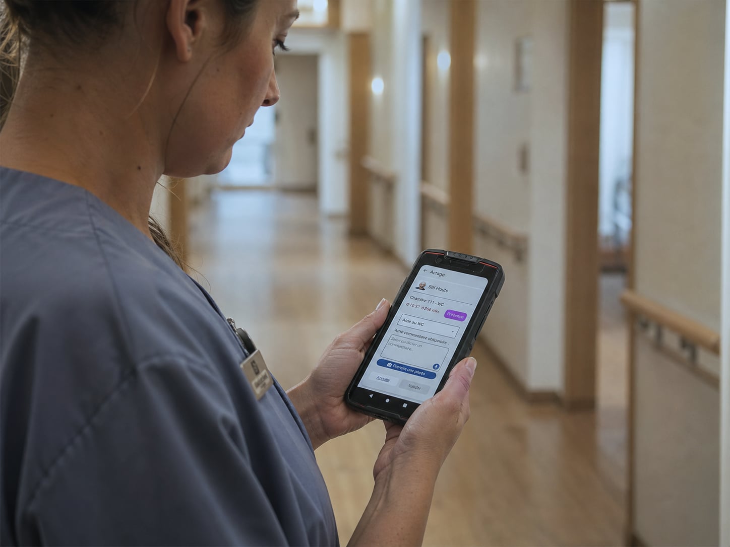 Soignante en polo bleu marine consultant une alerte d'appel infirmier sur un smartphone Android professionnel partagé par plusieurs membres du personnel, couloir d'une maison de repos belge équipée Healthcall